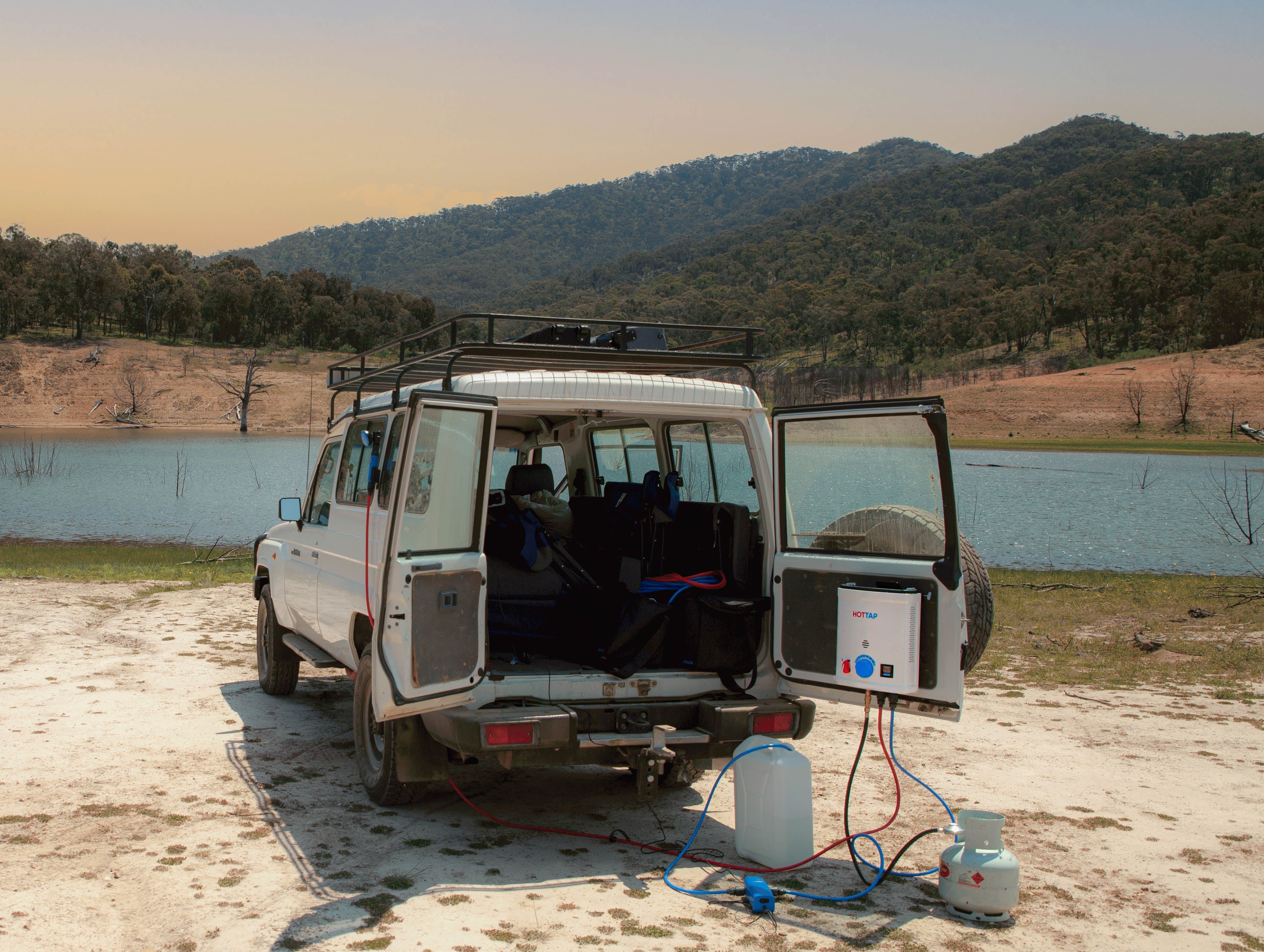 A 4WD in front of a large lake and hills with a portable water heater attached via the mounting bracket