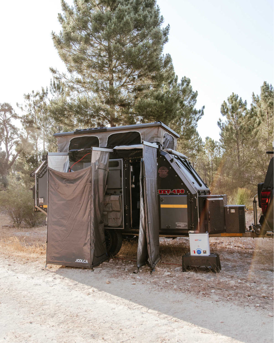 An open Joolca branded mounted double tent attached to a caravan out on a dirt road surrounded by trees