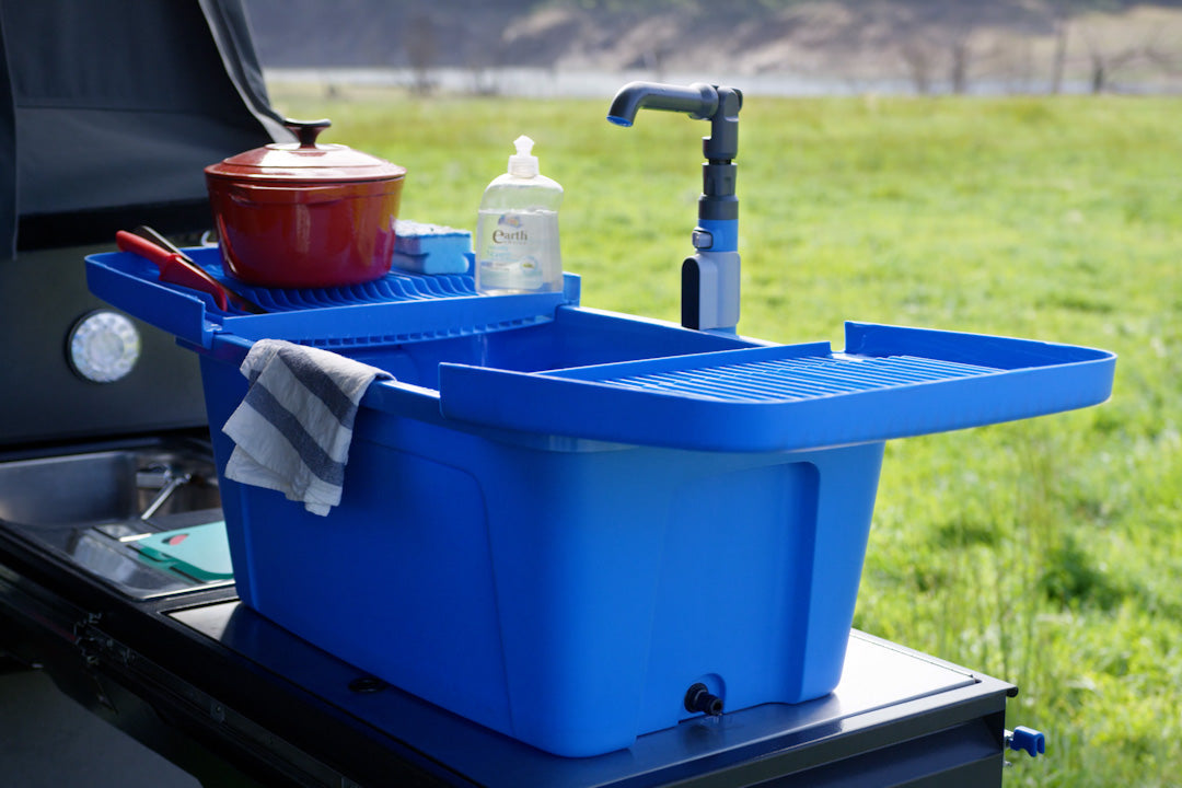 Blue Nomad Sink with its dish rack lid and faucet, set up on a camp kitchen with a pot and dish soap