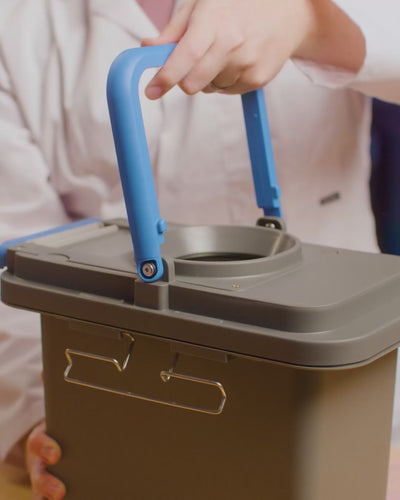 Hand lifting the GottaGo solids cassette by its blue folding handle, the bag opening visible in the lid.