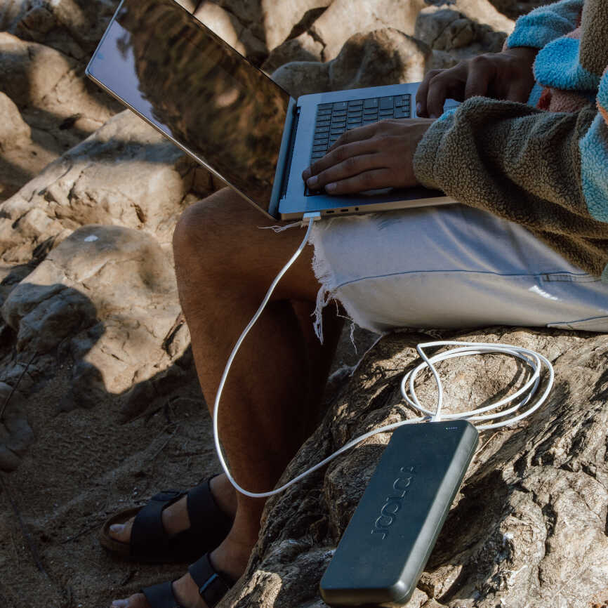 A man sitting on a rock using a laptop and charging the laptop via a power bank